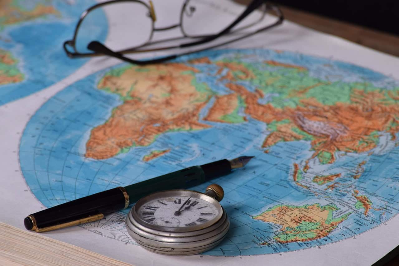 Pocket watch, fountain pen, and reading glasses resting on a colorful world map, suggesting travel planning.